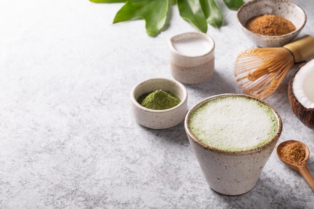 Zutaten für die Zubereitung von süßem Matcha Latte Japanischer grüner Tee mit Kokosmilch traditioneller Bambusbesen Chasen auf weißem Betonhintergrund Exemplar