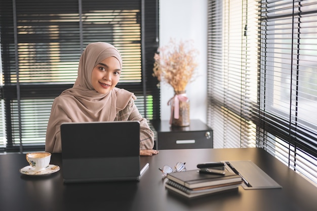 Zuversichtlich asiatische muslimische Geschäftsfrau brauner Hijab, der mit Laptop-Computer am modernen Büro sitzt und arbeitet.