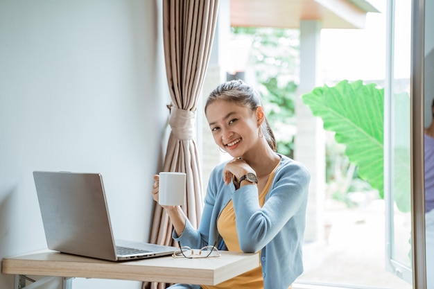 Zuversichtlich junge Frau in der intelligenten Freizeitkleidung, die am Laptop beim Sitzen nahe Fenster zu Hause arbeitet