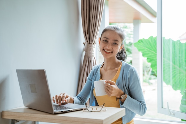 Zuversichtlich junge Frau in der intelligenten Freizeitkleidung, die am Laptop beim Sitzen nahe Fenster zu Hause arbeitet