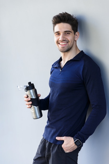 Zuversichtlich lächelnder Sportler, der Wasserflasche hält, während lokalisiert über grauem Hintergrund steht