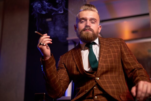 Zuversichtlich wohlhabender kaukasischer Mann mit Bart im formellen stilvollen Anzug sitzen rauchende Zigarre