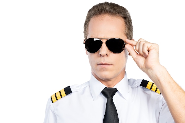 Zuversichtlicher Pilot. Selbstbewusster männlicher Pilot in Uniform, der seine Brille im Stehen vor weißem Hintergrund justiert