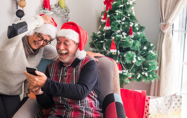 Zwei ältere Menschen im Ruhestand mit Santa's Cap mit Spaß am Handy suchen. Geschenke und Weihnachtsbaum im Hintergrund