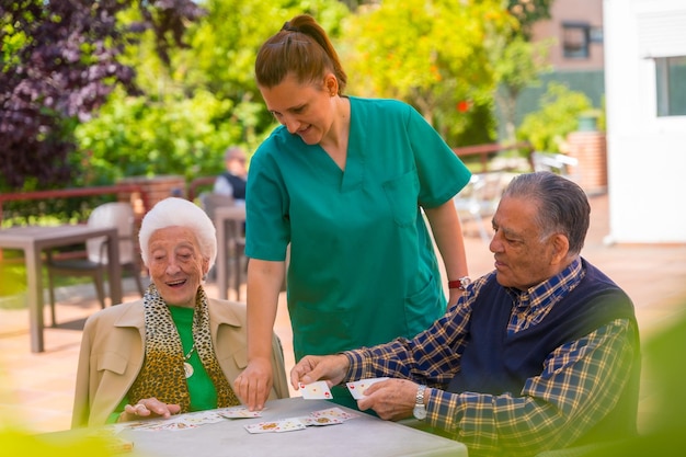 Zwei ältere Menschen spielen mit der Krankenschwester im Garten eines Pflegeheims oder Altersheims Karten