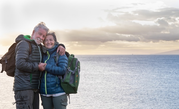 Zwei ältere Menschen umarmen sich vor dem Meer und genießen das Sonnenuntergangslicht Stehen auf der Klippe