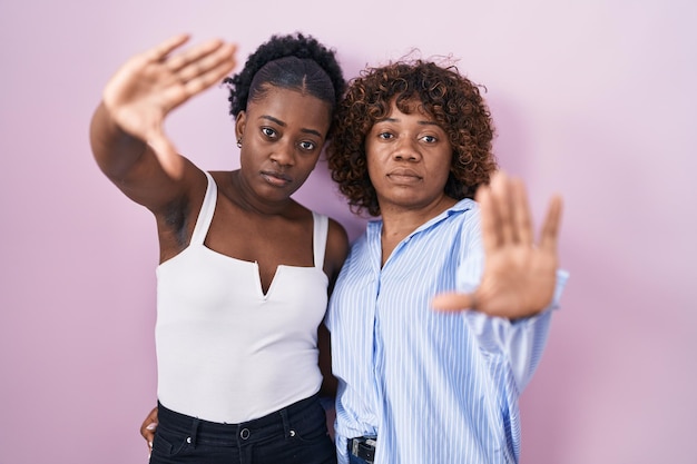 Zwei afrikanische Frauen stehen vor rosa Hintergrund und machen Bilder mit Händen, Handflächen und Fingern aus der Kameraperspektive