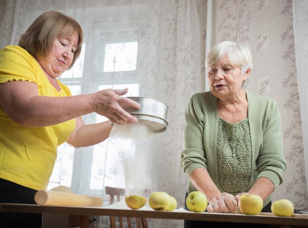 Zwei alte Frauen machen kleine Apfelkuchen zu Hause und sieben Mehl auf den Tisch