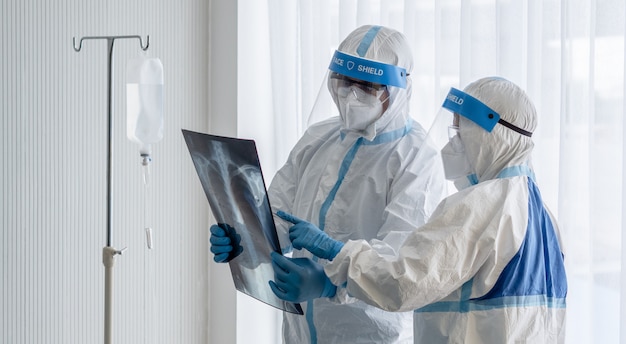 Zwei asiatische ärzte tragen einen psa-anzug mit n95-maske und gesichtsschutz und untersuchen den röntgenfilm der lungenbrust eines mit coronavirus infizierten patienten im unterdruckraum.