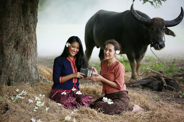 Zwei asiatische Frau, die traditionelles thailändisches Kostüm sitzt, sitzt im Feld, während Weinlesestilradio neben Büffel hört