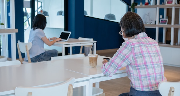 Zwei asiatische Frauen, die eine Gesichtsmaske tragen und Smartphone und Laptop für Videoanrufe oder zum Arbeiten verwenden, sitzen auf separaten Tischen, um die soziale Sicherheit als neues normales Lebensstilkonzept aufrechtzuerhalten.