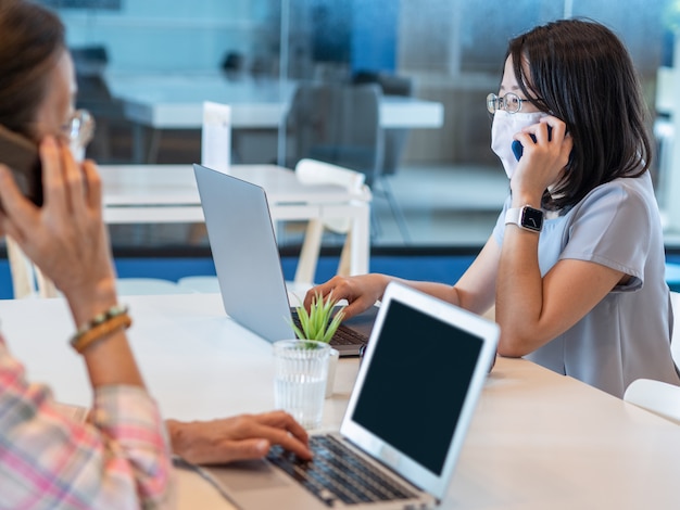 Zwei asiatische Frauen, die Gesichtsmaske tragen und Smartphone und Laptop für Videoanrufe oder Arbeiten verwenden