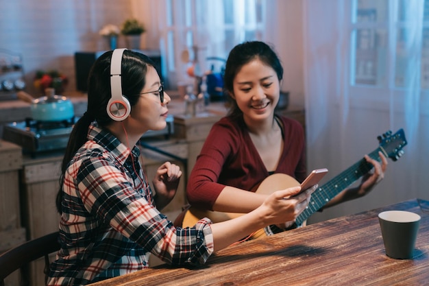 Zwei asiatische japanische Freundinnen sitzen um Mitternacht mit Gitarre in der modernen Holzküche zu Hause. junges Mädchen, das Noten auf dem Handy liest. frau in den kopfhörern, die handy halten, genießen melodie