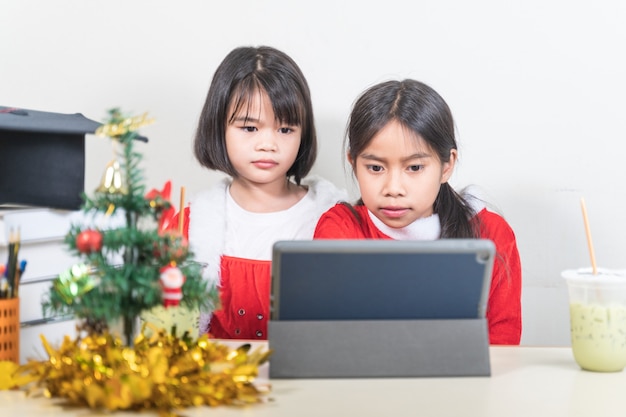 Zwei asiatische Kinder kleine Mädchen tragen Weihnachtsweihnachtsmann im Chat mit Freund auf digitalem Tablet. Weihnachtsferienkonzept Stockfotos