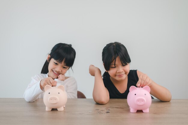 Zwei asiatische kleine Mädchen, die Spaß daran haben, zusammen Münzen in Piggy Bank zu stecken, Kinder sparen Geld für das zukünftige Konzept