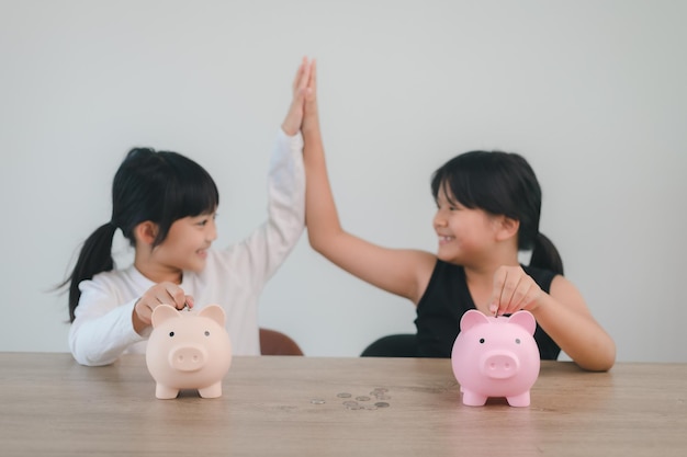 Zwei asiatische kleine Mädchen, die Spaß daran haben, zusammen Münzen in Piggy Bank zu stecken, Kinder sparen Geld für das zukünftige Konzept