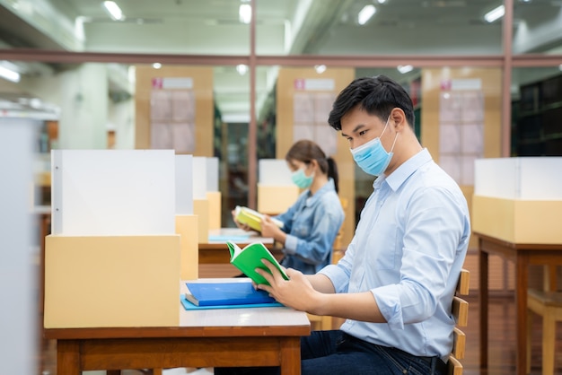 Zwei asiatische Studenten, die Gesichtsmaske tragen und in der sozialen Entfernung der Bibliothek von anderen 6 Fuß sitzen