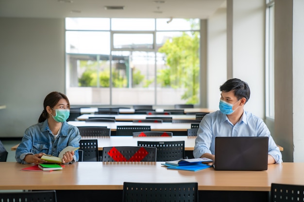 Zwei asiatische Universitätsstudenten, die Gesichtsmaske tragen und in der sozialen Entfernung der Bibliothek von anderen sitzen