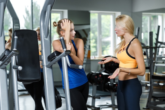 Zwei athletische blonde Frauen, die im Fitnessstudio sprechen, kommunizieren mit Trainer