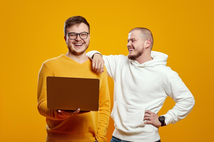 Zwei attraktive Freunde, Männer mit gelbem Pullover und weißem Hoodie, lachen, während sie einen Laptop halten
