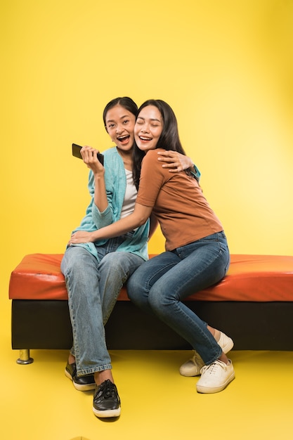 Zwei attraktive junge Frauen lachen und umarmen sich beim scherzenden Sitzen und eine hält ein Handy
