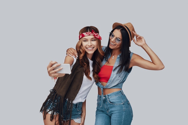 Zwei attraktive, stilvolle junge Frauen, die Selfie machen und lächeln, während sie vor grauem Hintergrund stehen