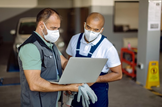 Zwei Automechaniker tragen Gesichtsmasken, während sie in einer Werkstatt am Laptop arbeiten