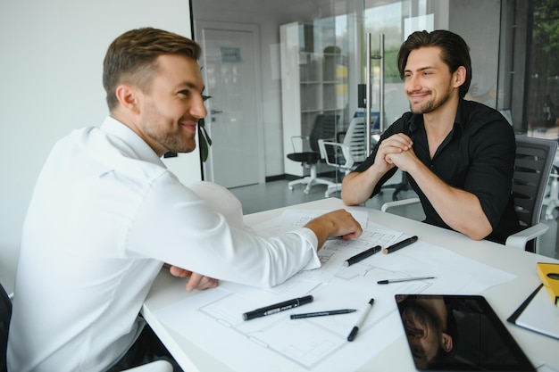 Zwei Bauplaner stehen in einem modernen Büro und lehnen sich über einen Schreibtisch, um gemeinsam Baupläne zu diskutieren