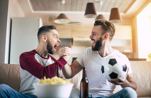 Zwei beste Freunde und Fußballfans schauen sich ein Sportspiel im Fernsehen an, trinken Bier und essen Snacks, während sie das Team auf der Couch anfeuern