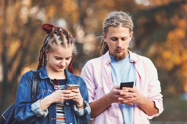 Zwei beste Hipster-Freunde mit Dreads, die lässige Kleidung tragen, Nachrichten auf ihren Smartphones eingeben und sich im Freien nicht gegenseitig beachten