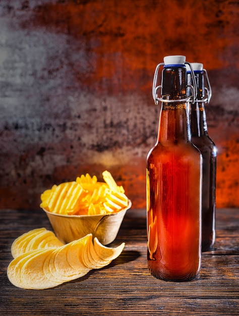 Zwei Bierflaschen in der Nähe von verstreuten Pommes und einem Teller mit Snacks auf einem dunklen Holzschreibtisch. Lebensmittel- und Getränkekonzept