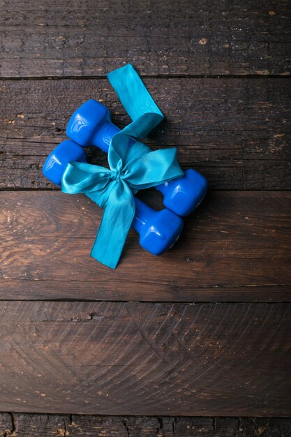Zwei blaue Hantel mit einer Geschenkschleife und einem leeren Papieranhänger, Herz auf Holzbrettern, Sportferienkonzept