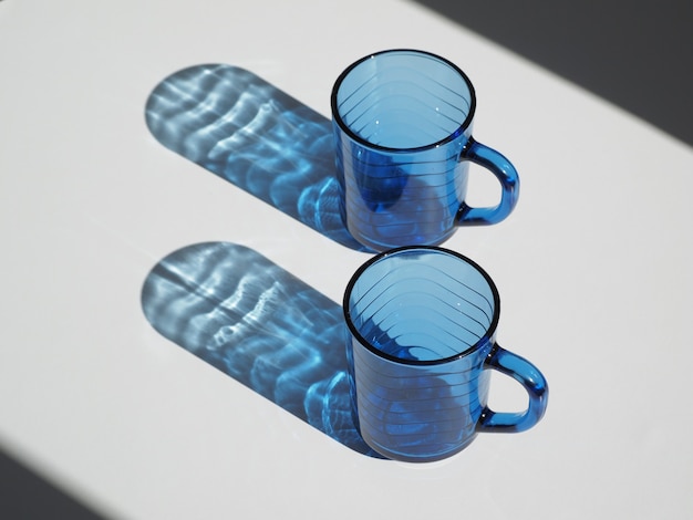 Zwei blaue leere Gläser mit Sonnenlichtreflexion auf Weiß. Platz für Text.