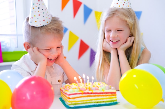 Zwei blonde kaukasische Kinder Jungen und Mädchen haben Spaß beim Ausblasen von Kerzen beim Geburtstagsregenbogenkuchen mit cake