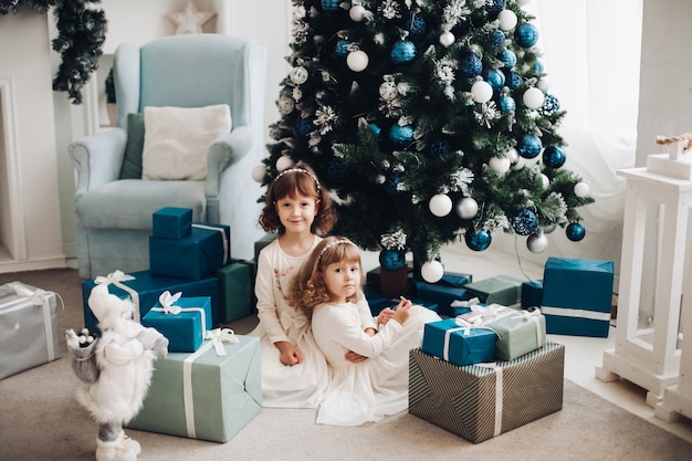 Zwei blondhaarige kleine Mädchen in weißen Kleidern sitzen nahe Geschenken unter Weihnachtsbaum