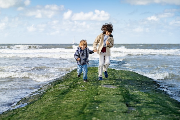 Zwei Brüder laufen Händchen haltend auf einem Wellenbrecher an der Nordsee. Lachende Jungs verbringen ein Wochenende am Meer in Belgien, Knokke. Geschwisterfreundschaft.