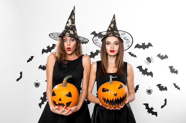 Zwei brünette Mädchen in schwarzen Kleidern und Hexenhüten halten Horror-Halloween-Kürbisse mit Fledermäusen im Hintergrund.