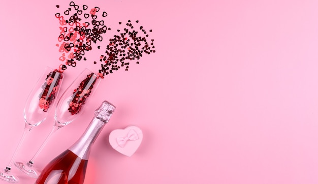 Foto zwei champagnergläser gefüllt mit herzförmigen pailletten mit einer flasche champagner und einem valentinstaggeschenk auf einem rosa hintergrund.