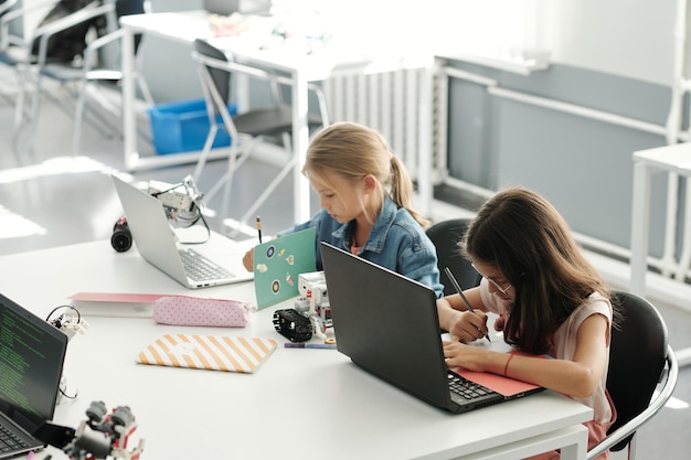 Zwei clevere Schulmädchen machen sich vor Laptops Notizen in ihren Schulheften