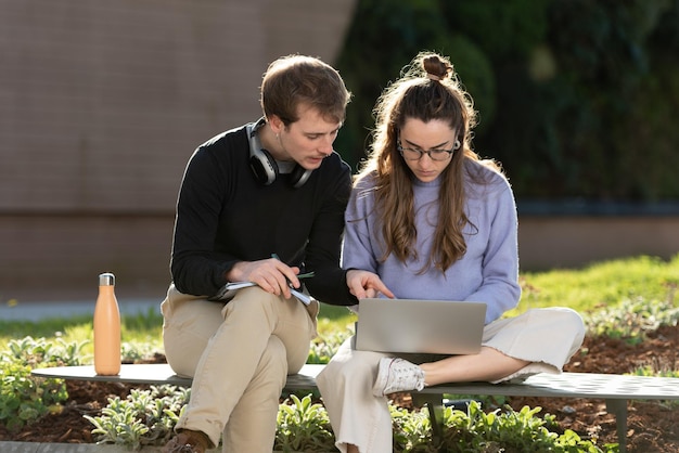 Zwei College-Studenten sitzen zusammen auf einer Bank im Freien. Junges Paar, das an einem Laptop in einem Park arbeitet.