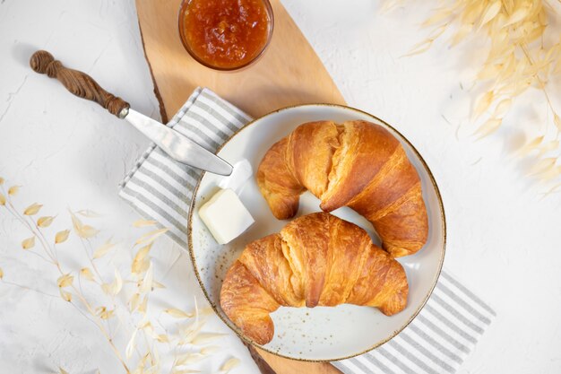 Zwei Croissants auf Keramikplatte mit Butter und Orangenmarmelade auf einem Holzbrett. Draufsicht.