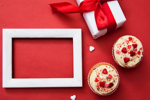 Zwei Cupcakes mit Sahne und Dekor, Geschenk, weißer Rahmen auf rotem Hintergrund. Flache Zusammensetzung des glücklichen Valentinstags