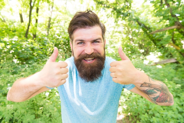 Foto zwei daumen hoch für den sommer. hipster, der auf die natur gestikuliert. kaukasischer hipster mit dickem barthaar und stilvollem haarschnitt. brutaler hipster auf naturlandschaft. bärtiger mann im trendigen hipster-stil im freien.