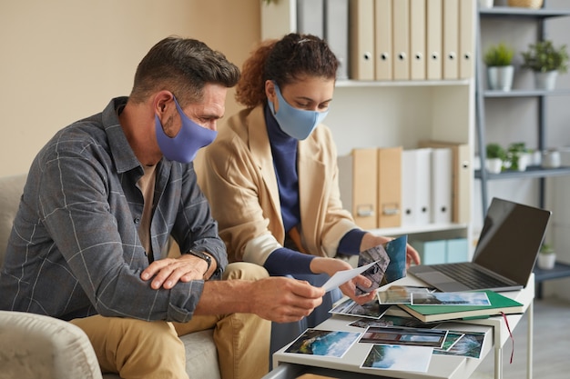 Zwei Designer in Schutzmasken arbeiten während ihres Treffens im Büro mit professionellen Fotos im Team