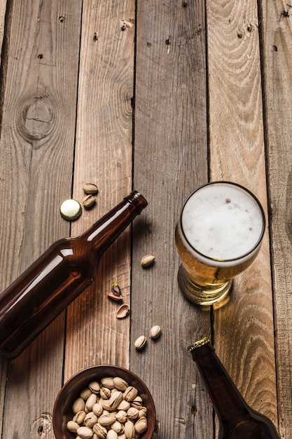 Zwei dunkle Bierflaschen, ein Glas Bier und Schaum und ein Teller Pistazien auf einem hölzernen Hintergrund.