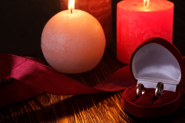 Zwei Eheringe in der roten Geschenkbox mit Kerzenlicht mit drei Wachsflammen mit Band im dunklen romantischen Licht, Liebesdatierung, Valentinstag, selektiver Fokus