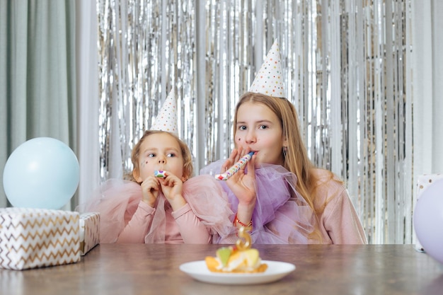 Zwei einjährige Schwestern mit Partyhüten und eleganten Kleidern sitzen am Tisch und blasen in die Luft