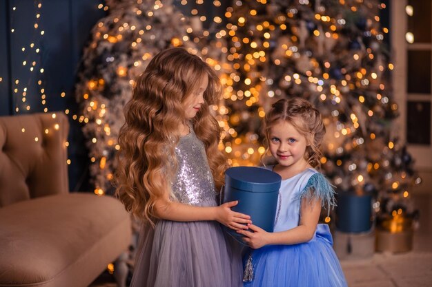 Zwei elegante Mädchen in schönen Kleidern beschenken sich gegenseitig vor dem Hintergrund eines Weihnachtsbaums mit Lichtern