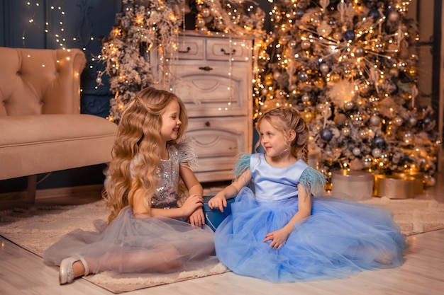 Zwei elegante Mädchen in schönen Kleidern beschenken sich gegenseitig vor dem Hintergrund eines Weihnachtsbaums mit Lichtern