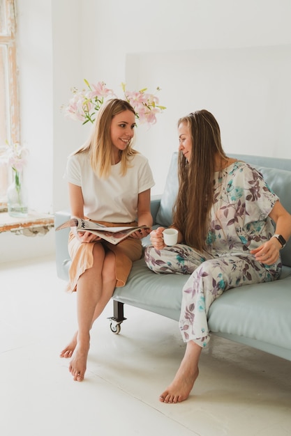 Zwei entzückende Frauen unterhalten sich zu Hause auf dem Sofa, trinken Kaffee und schauen sich ein Modemagazin an